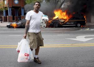 A man with goods looted from a store walks past burning vehicles during clashes in Baltimore, Maryland